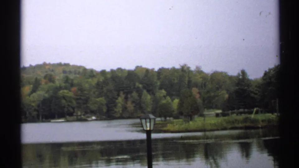 1971 New York 8mm frame showing calm lake water, wooded shoreline, early autumn foliage, and a dark foreground lamppost.