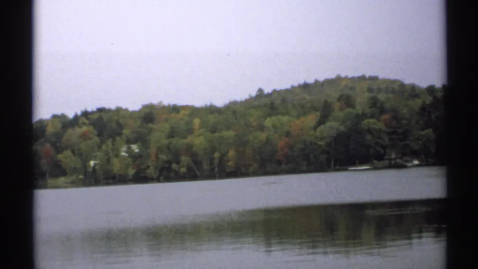 1971 New York 8mm frame looking across still lake water toward a tree-covered hill with muted autumn color.