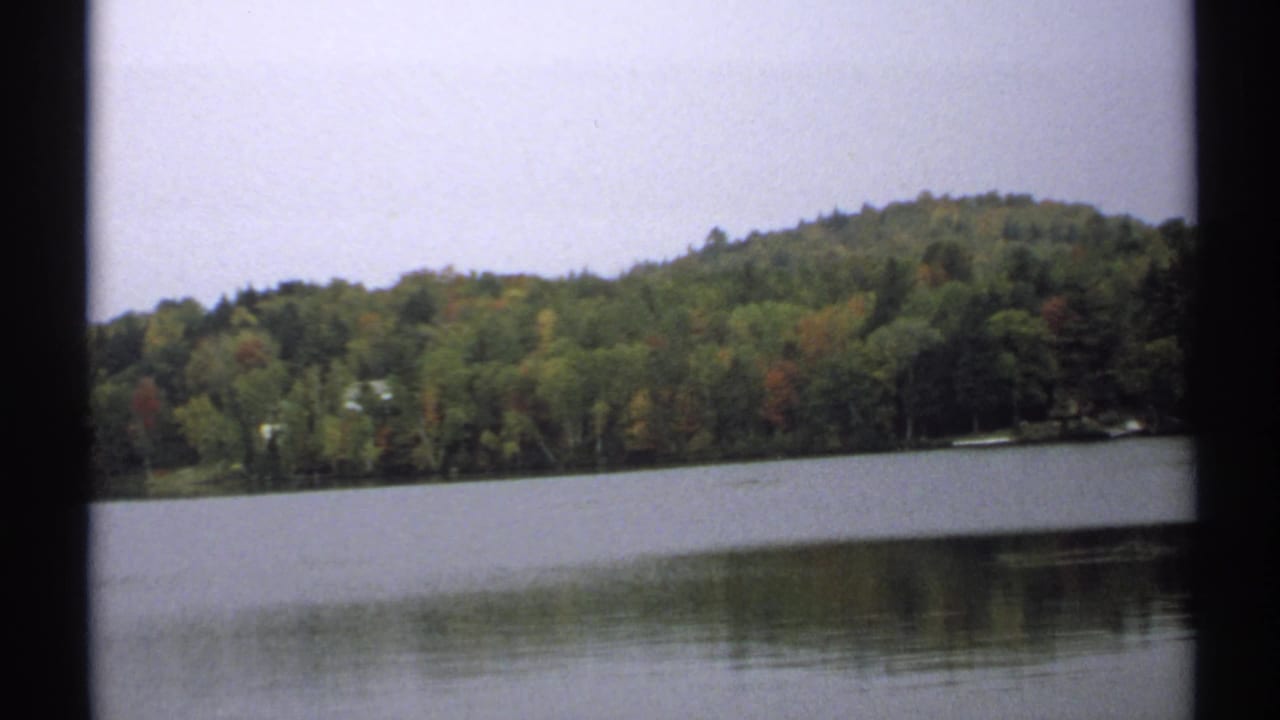 1971 New York home-movie frame showing a lake, distant wooded slope, and subdued green, yellow, and red foliage.
