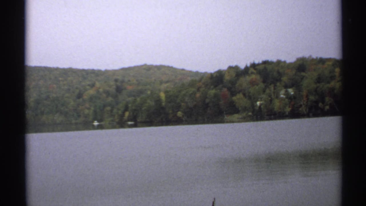 1971 New York 8mm frame of calm lake water facing a wooded shoreline and low hills under a pale sky.