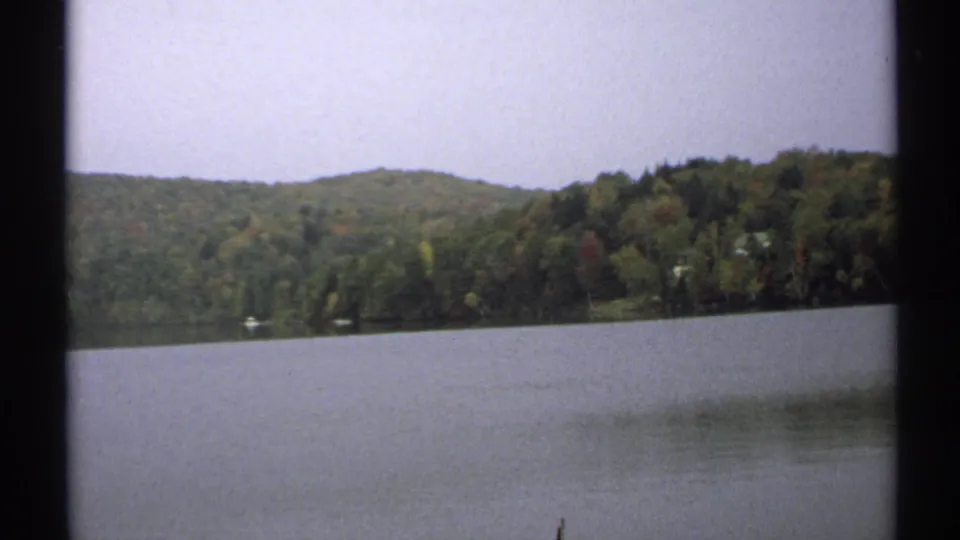 1971 New York 8mm frame of calm lake water facing a wooded shoreline and low hills under a pale sky.