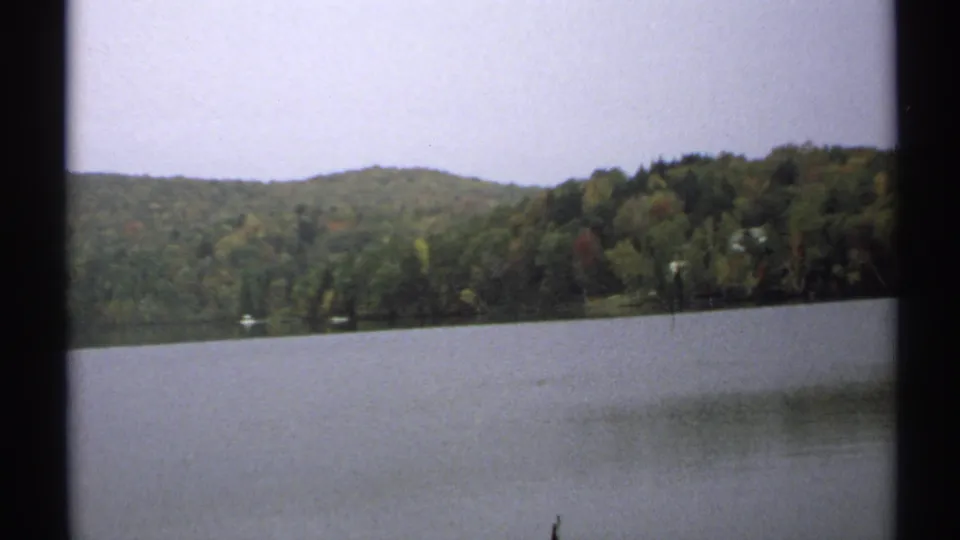 Wide 1971 New York lakeside frame with still water, wooded hills, faint boats near shore, and visible 8mm contact-frame borders.