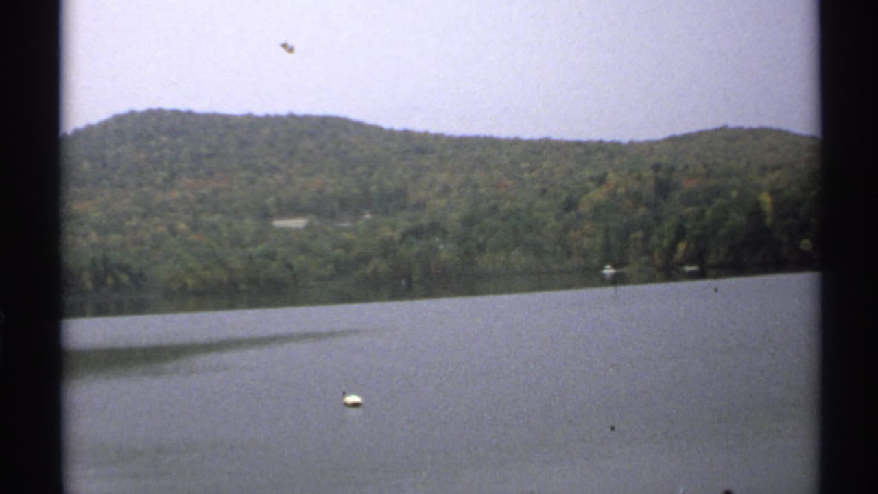 1971 New York home-movie frame showing calm lake water, a small white buoy, wooded hills, and early autumn color in soft 8mm texture.
