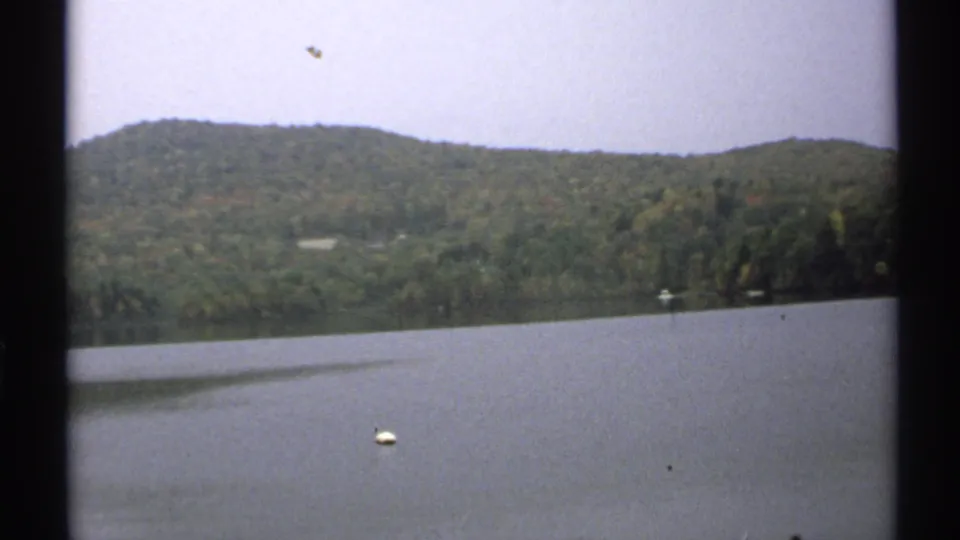 1971 New York home-movie frame showing calm lake water, a small white buoy, wooded hills, and early autumn color in soft 8mm texture.