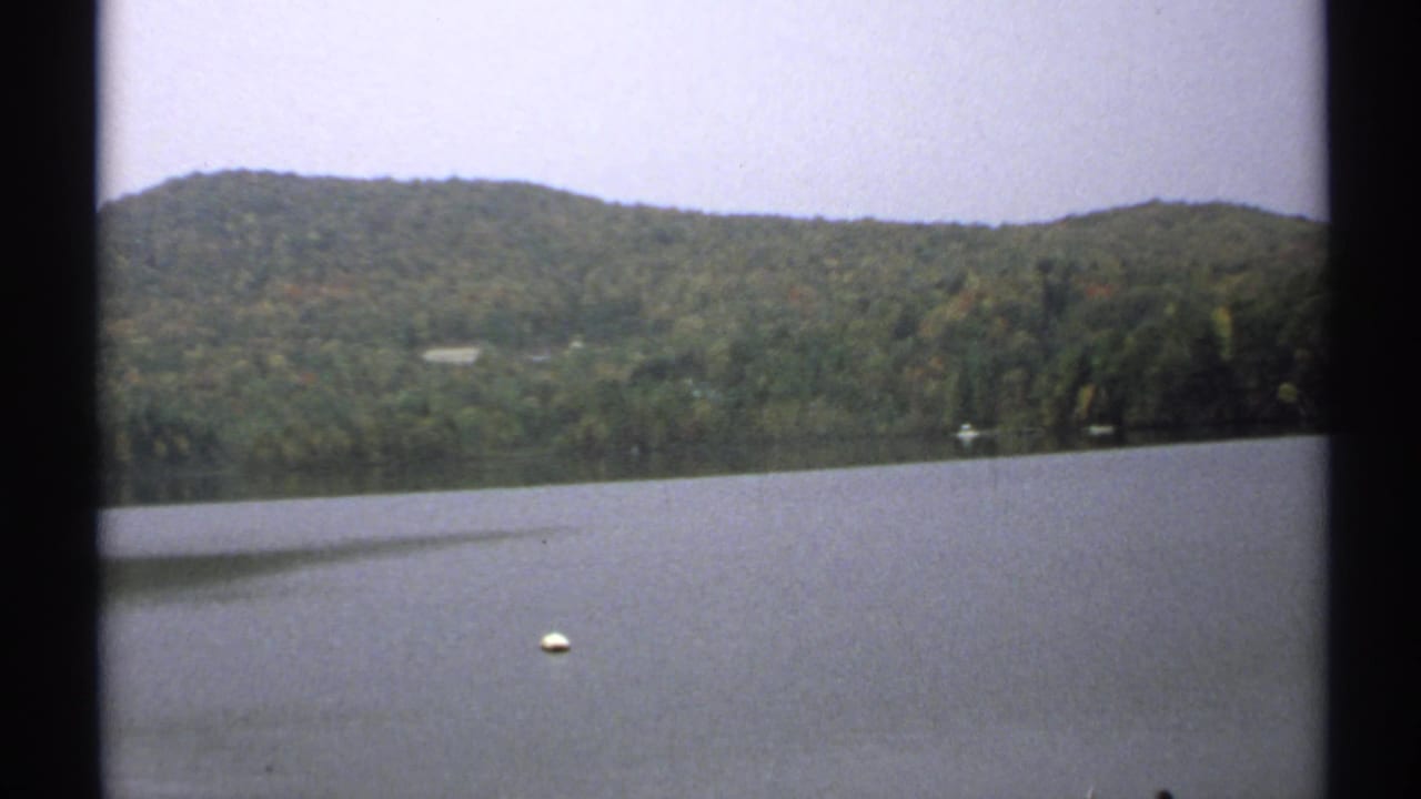 Quiet 1971 New York lake scene with a white buoy in the foreground, wooded hills beyond the water, and soft grain from the 8mm source.
