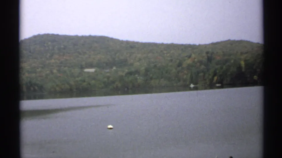 Quiet 1971 New York lake scene with a white buoy in the foreground, wooded hills beyond the water, and soft grain from the 8mm source.