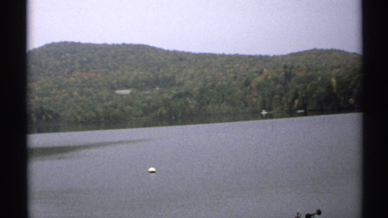 Calm lake with a wooded hill, pale sky, and white buoy in the foreground.