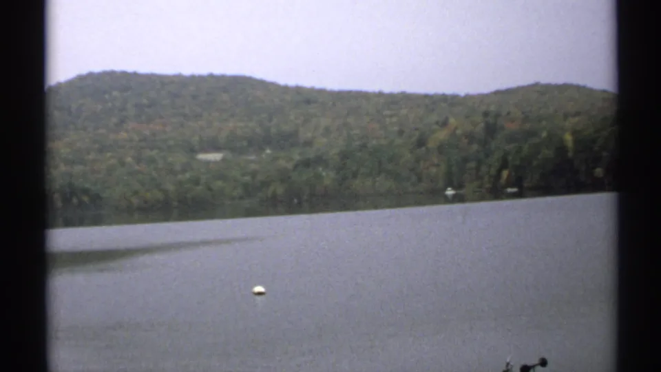 1971 New York 8mm frame showing a calm lake, tree-covered hill, overcast sky, and a small white buoy.