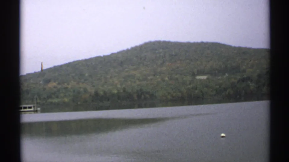 1971 New York lake view with forested hills, still water, a distant dock or structure, and a white buoy.