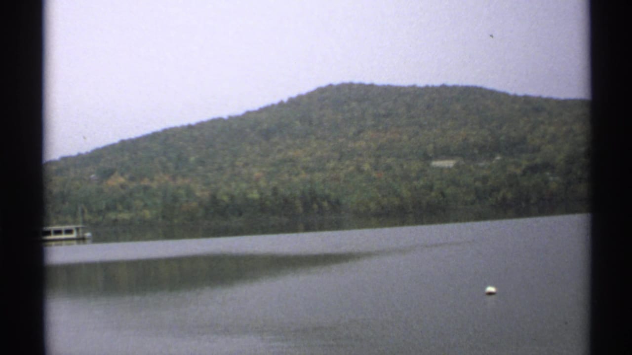 1971 New York 8mm frame of a calm lake, wooded hillside, partial lakeside dock, and floating white buoy.