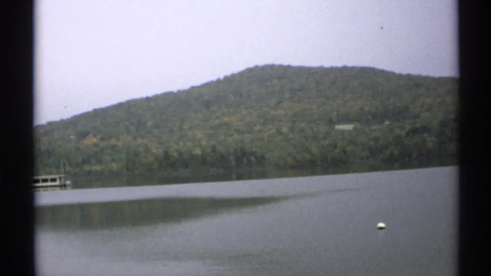 1971 New York 8mm frame of a calm lake, wooded hillside, partial lakeside dock, and floating white buoy.