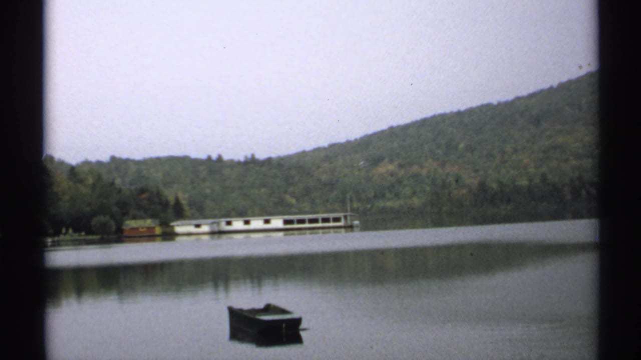 1971 New York lake scene with a small dark boat in the foreground, still water, wooded hill, and distant dock or structure.