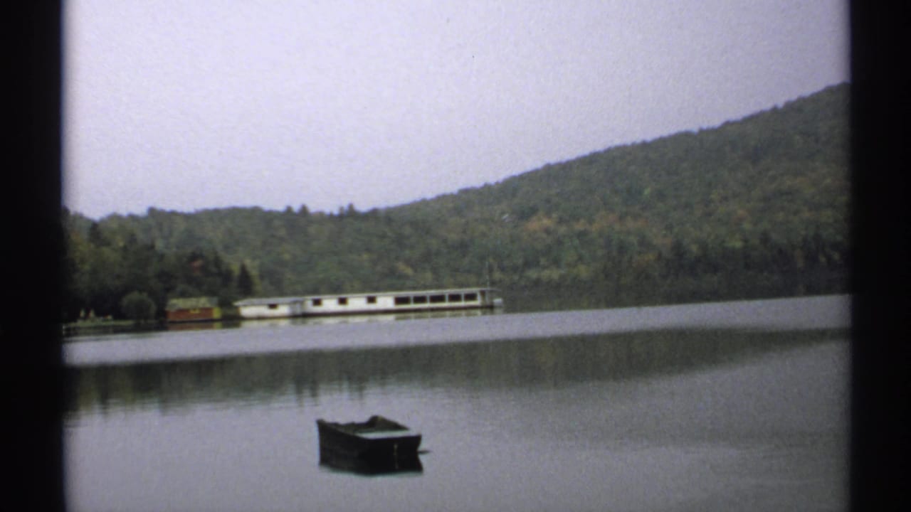 1971 New York 8mm frame showing a rowboat on calm lake water, with forested hills and a long white boathouse on the far shore.