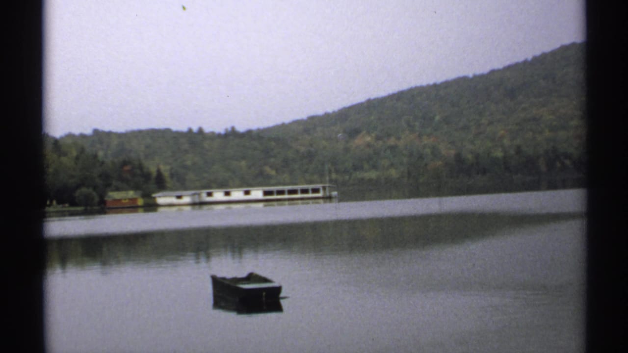 1971 New York 8mm view of a dark rowboat on a quiet lake, backed by wooded hills and a long white boathouse or lakeside structure.
