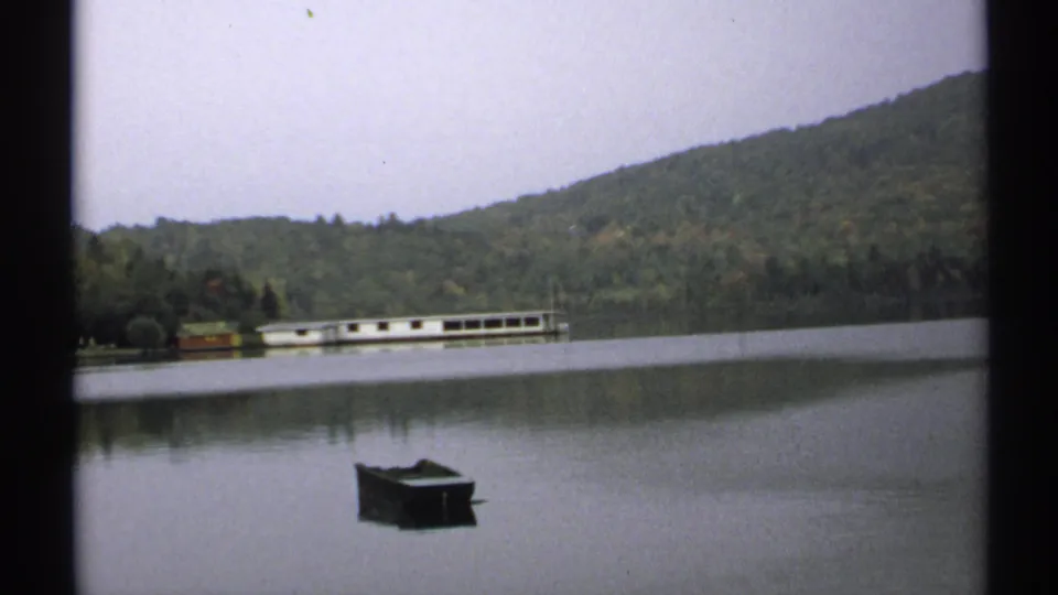 1971 New York 8mm view of a dark rowboat on a quiet lake, backed by wooded hills and a long white boathouse or lakeside structure.