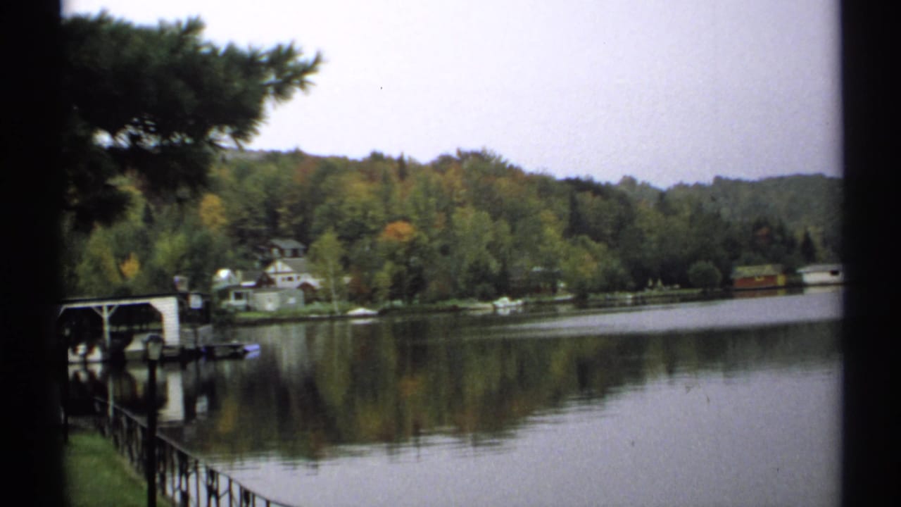 1971 New York 8mm frame showing a quiet lakeshore with a dock or boat shelter, small houses, wooded trees, and reflections on the water.
