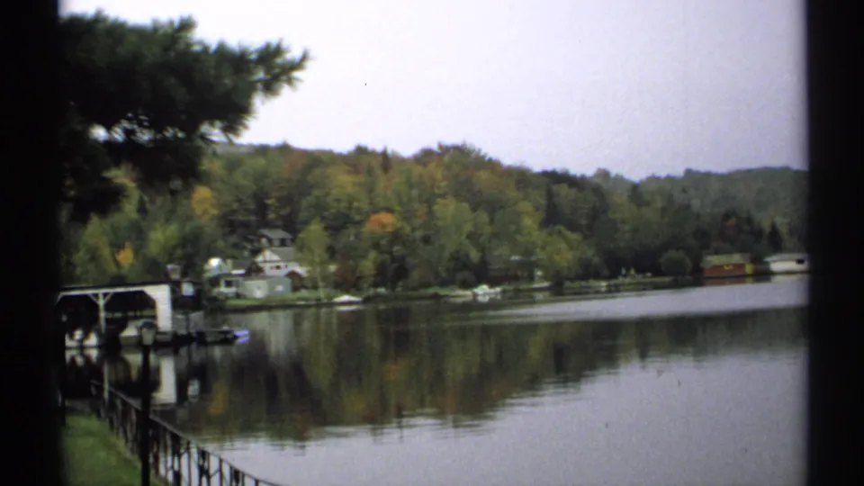 1971 New York 8mm frame showing a quiet lakeshore with a dock or boat shelter, small houses, wooded trees, and reflections on the water.