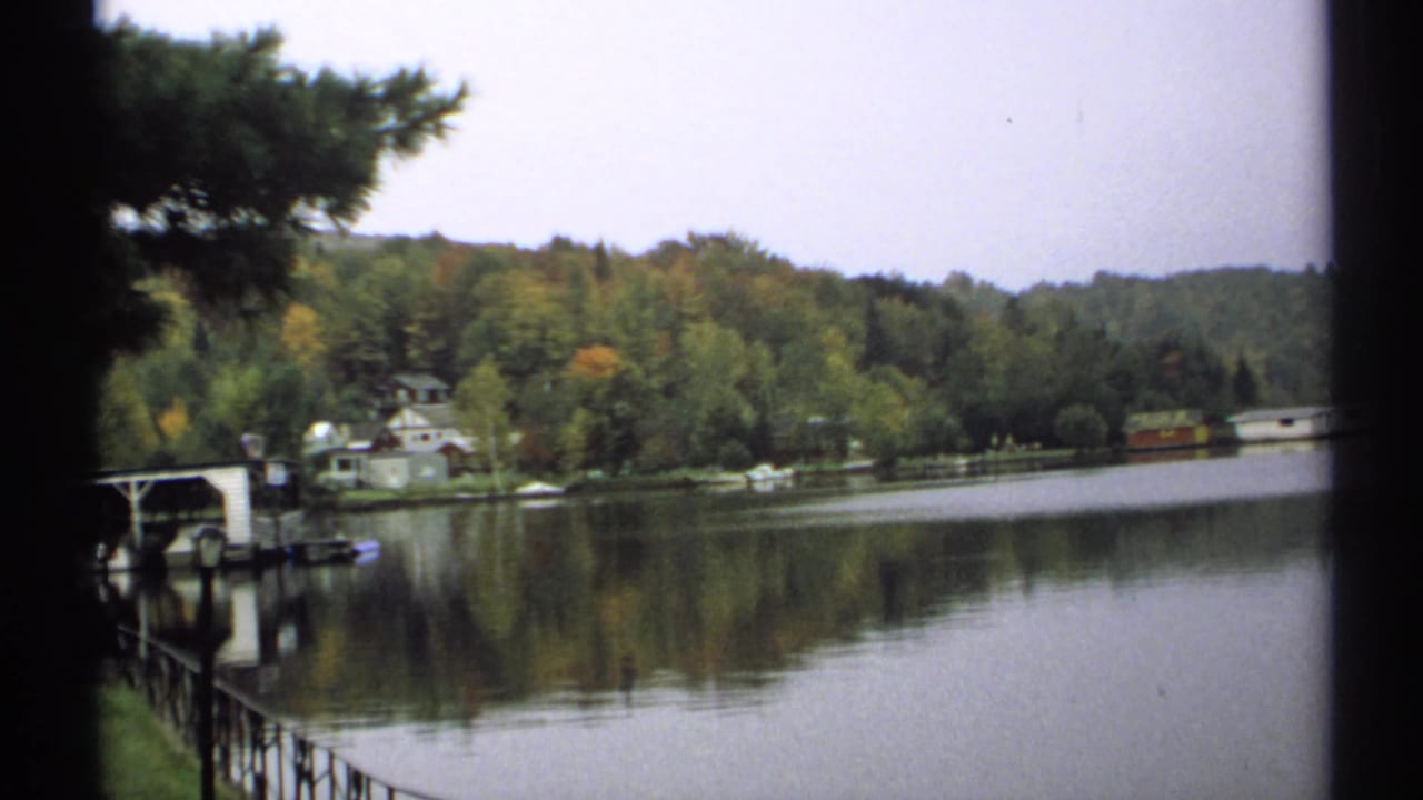 1971 New York 8mm lakeside view with shoreline homes, a dock or boat shelter, dense trees, reflective water, and a distant boathouse.