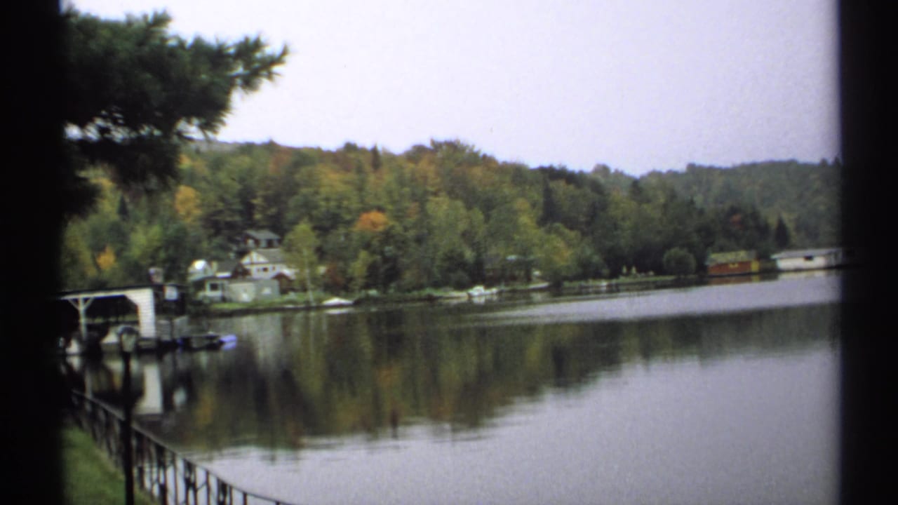 Calm lake with wooded shoreline, small houses, and a dark film edge at left.