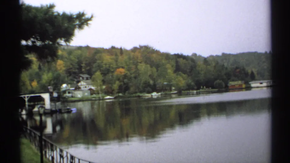 1971 New York 8mm frame of a calm lake, autumn trees, shoreline houses, and a partial dock or boathouse at the left edge.
