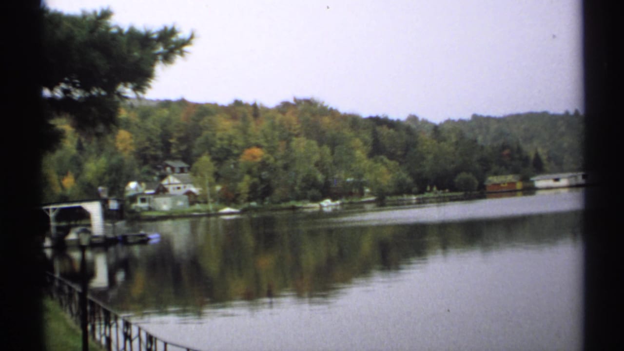 1971 New York 8mm view across a quiet lake toward trees, shoreline buildings, and a small structure near the water.