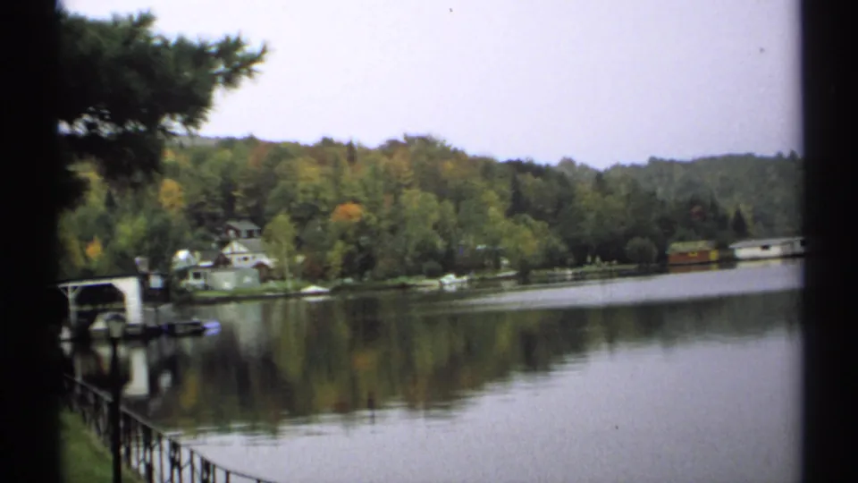 1971 New York 8mm view across a quiet lake toward trees, shoreline buildings, and a small structure near the water.