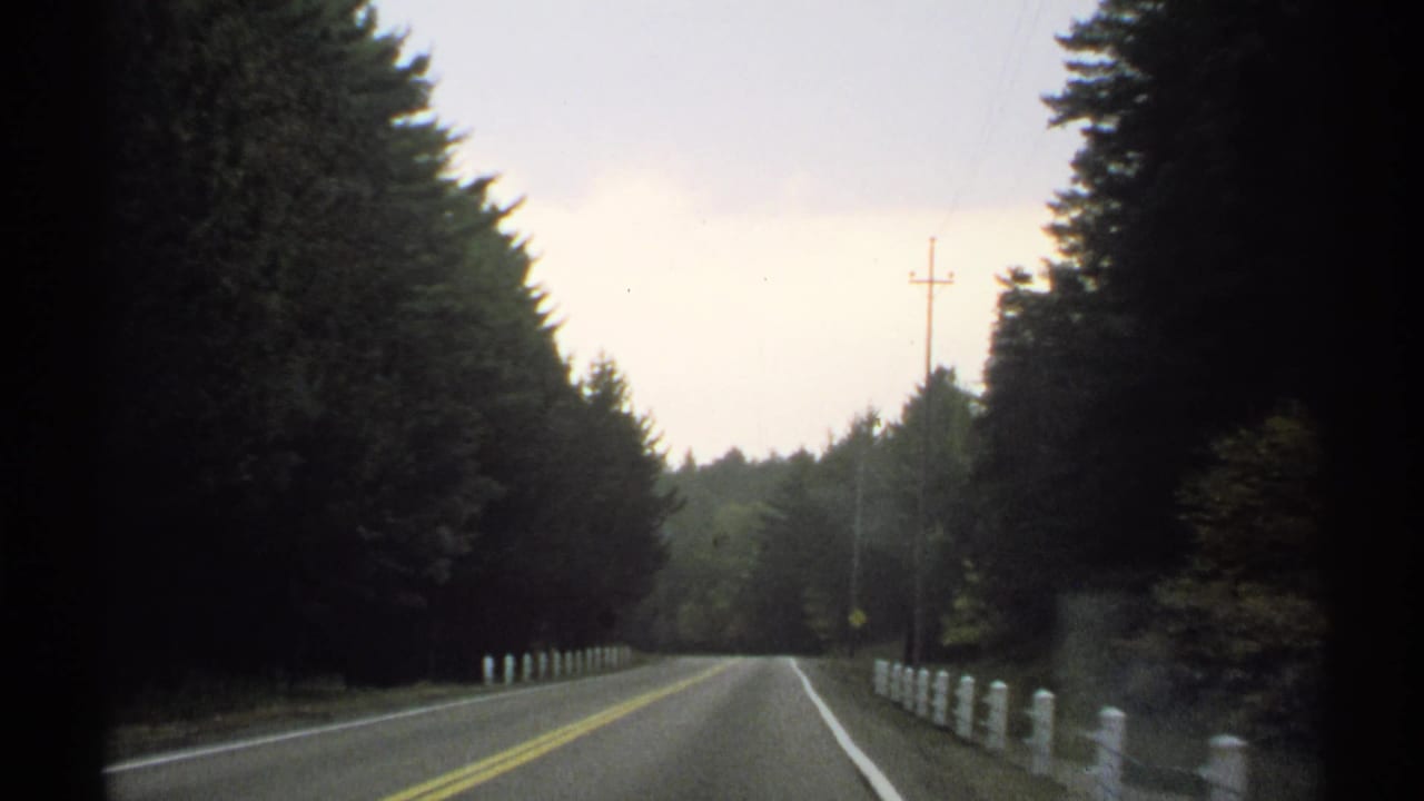 1971 New York 8mm frame of an empty two-lane road bordered by trees, white posts, and utility poles, filmed from a moving car.