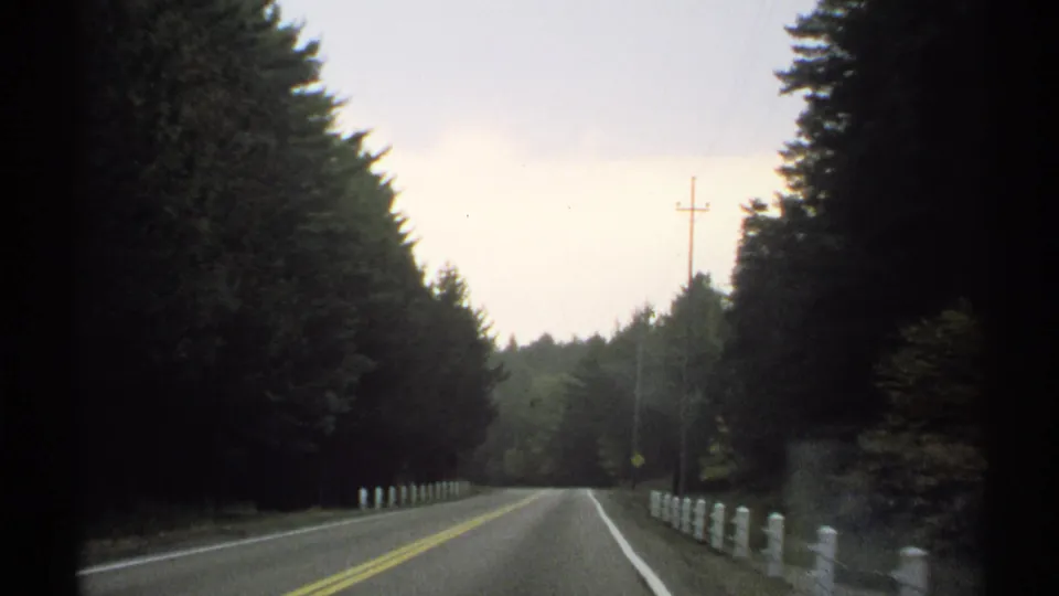 1971 New York 8mm frame of an empty two-lane road bordered by trees, white posts, and utility poles, filmed from a moving car.