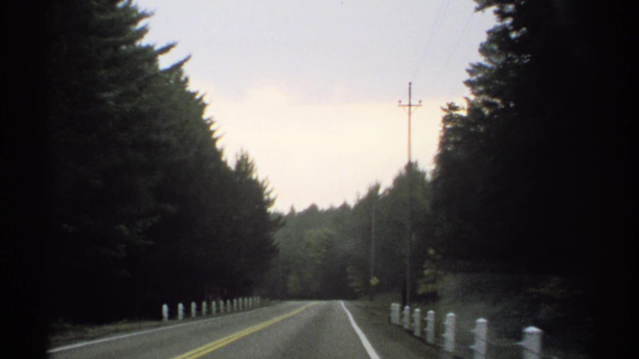 1971 New York 8mm frame looking down a quiet two-lane road bordered by dense evergreens and white roadside posts.