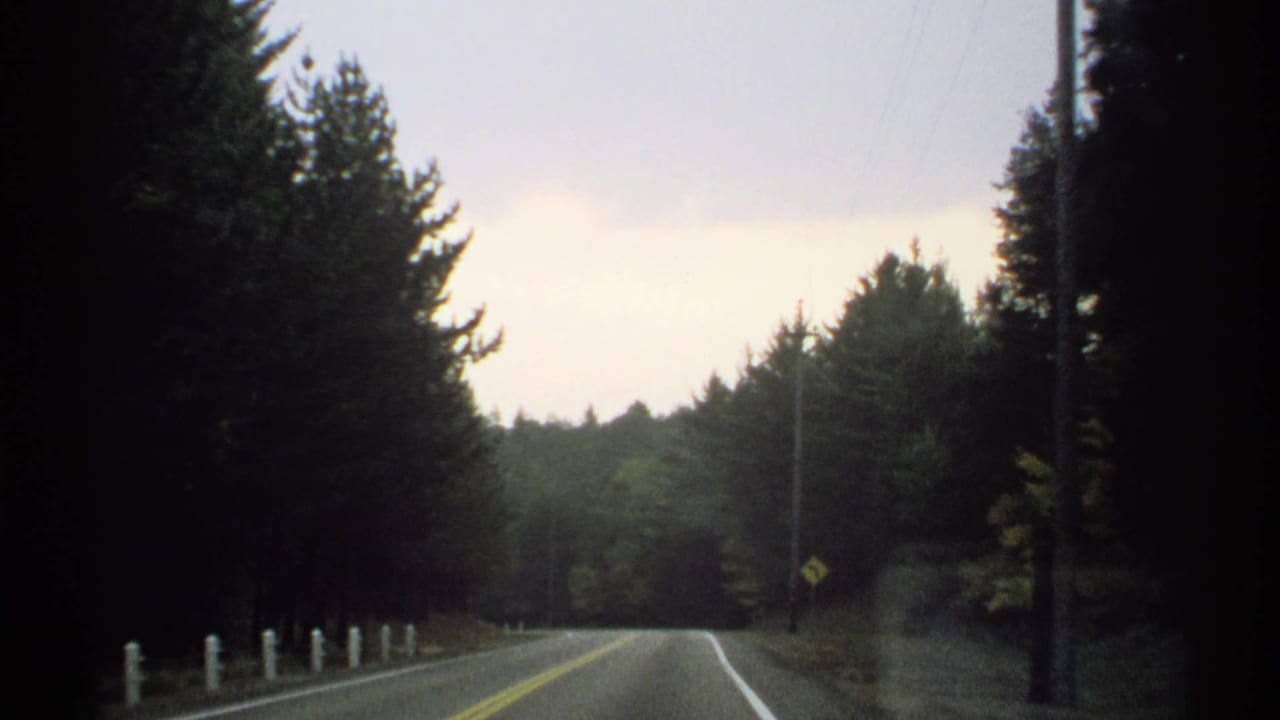 1971 home-movie frame from a moving car on a wooded New York road with white posts and a roadside utility pole.