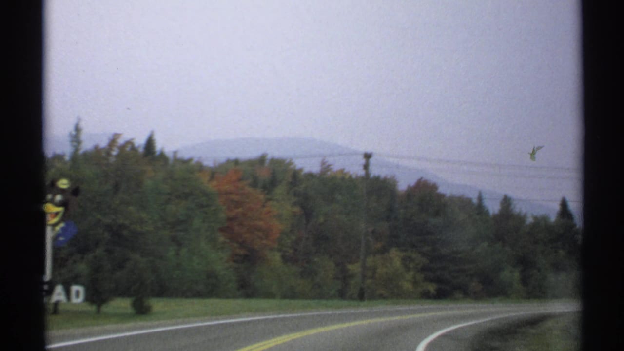 The road curves past trees toward distant New York hills, with muted 8mm color and traces of autumn foliage.