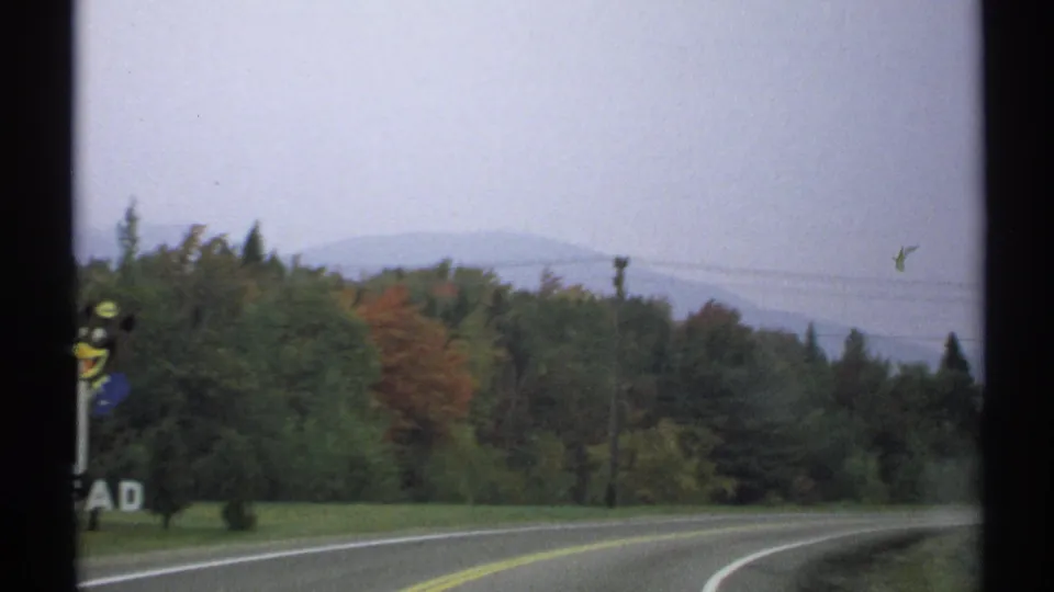 The road curves past trees toward distant New York hills, with muted 8mm color and traces of autumn foliage.
