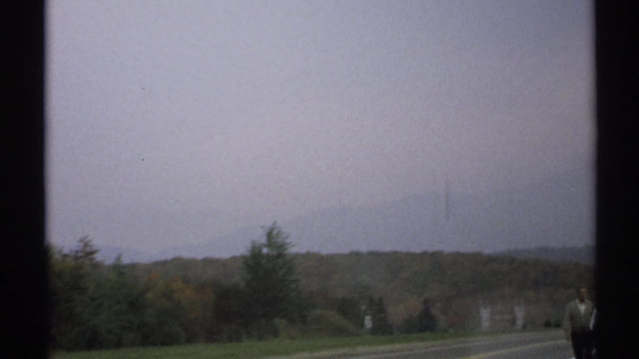 1971 rural New York road view with wooded hills, hazy sky, and a distant person partially visible near the right edge.