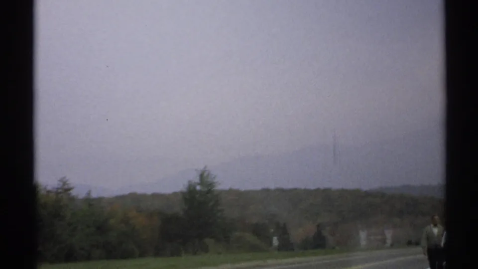 1971 rural New York road view with wooded hills, hazy sky, and a distant person partially visible near the right edge.