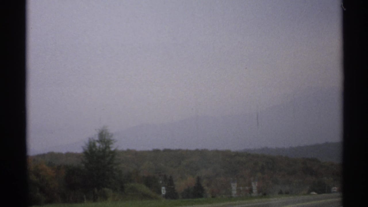 1971 upstate New York road view with autumn hills, distant mountains, and an overcast sky seen from a moving vehicle.
