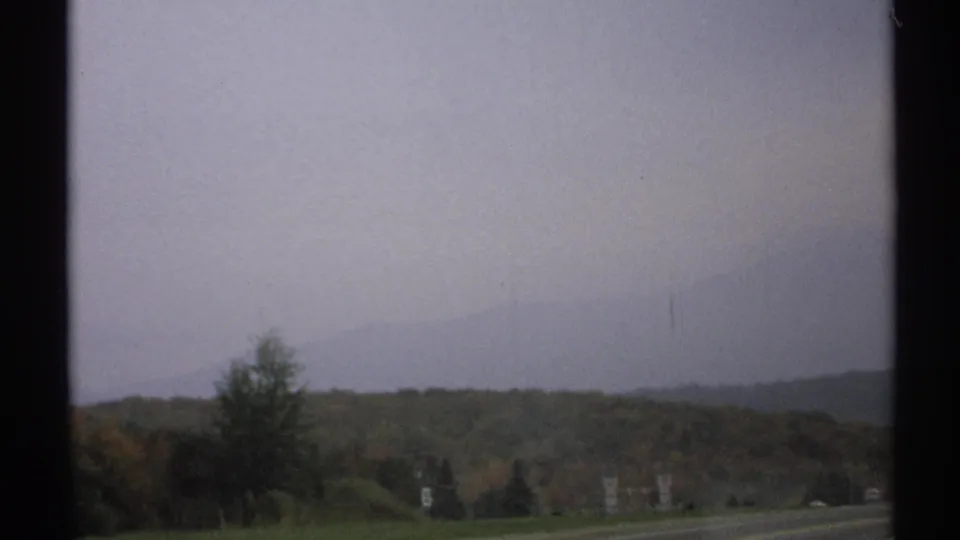 1971 upstate New York road view with autumn hills, distant mountains, and an overcast sky seen from a moving vehicle.