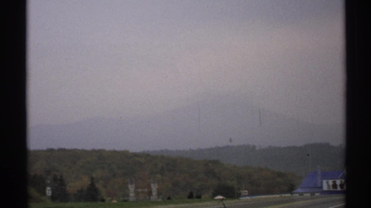 Upstate New York roadside scene with autumn hills, a blue-roofed building, and signs near the approach to Lake Placid.