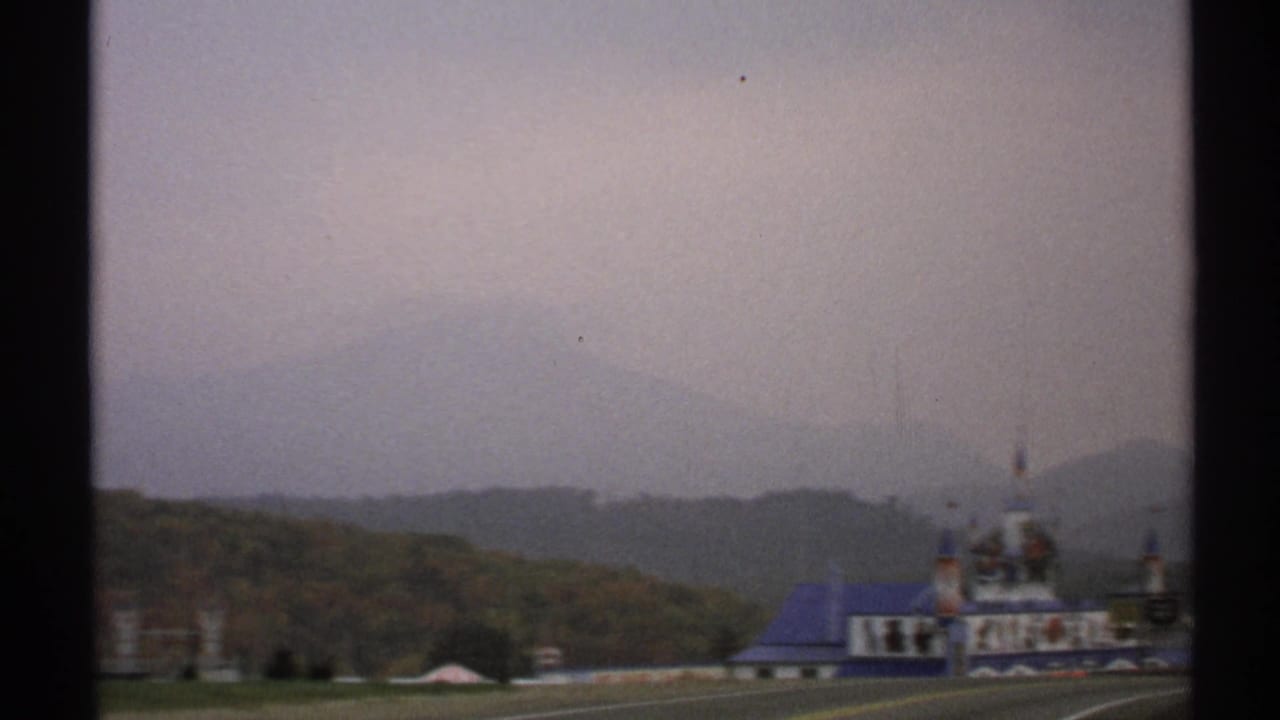 1971 home-movie frame showing a blue-roofed roadside structure beneath hazy hills in the Lake Placid area.