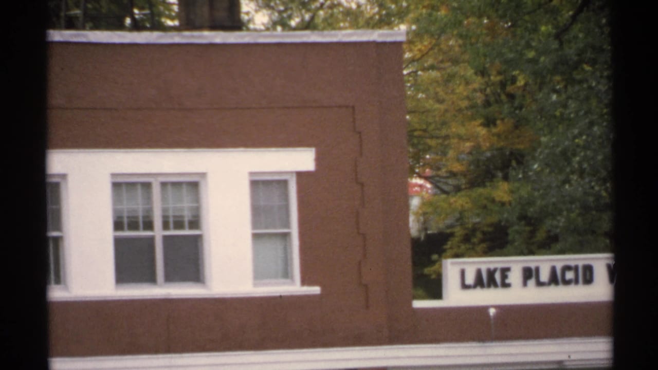 Lake Placid roadside sign beside a brown building with white-trimmed windows and autumn trees in the background.