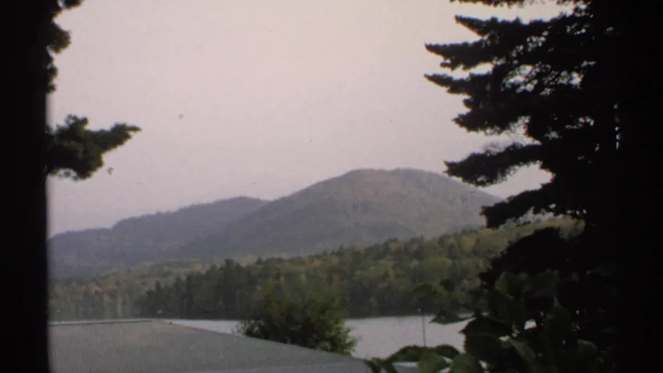 1971 New York 8mm scenic view of a lake, wooded hills, and pine branches under overcast light.