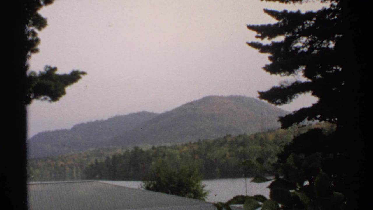 1971 New York 8mm landscape frame with wooded mountain slopes, lake water, pine branches, and muted sky.