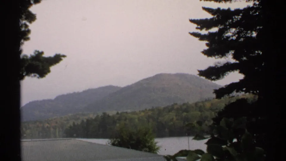1971 New York 8mm landscape frame with wooded mountain slopes, lake water, pine branches, and muted sky.