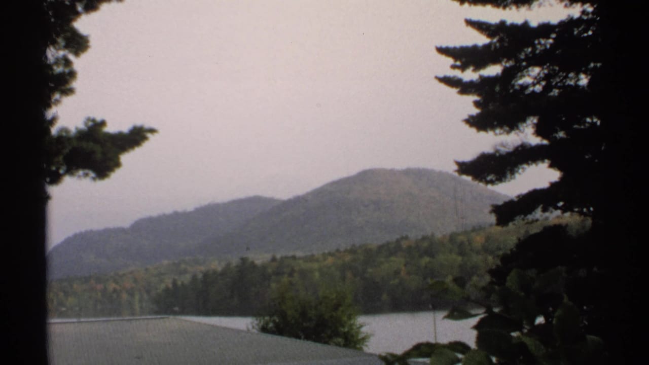 1971 New York 8mm view across a lake toward forested hills, with tree branches and foliage at the frame edge.