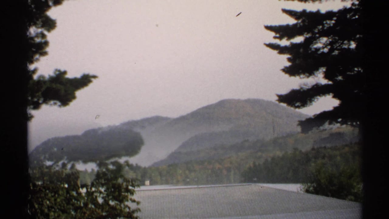 1971 New York 8mm frame showing a calm lake and forested mountains partly framed by trees.