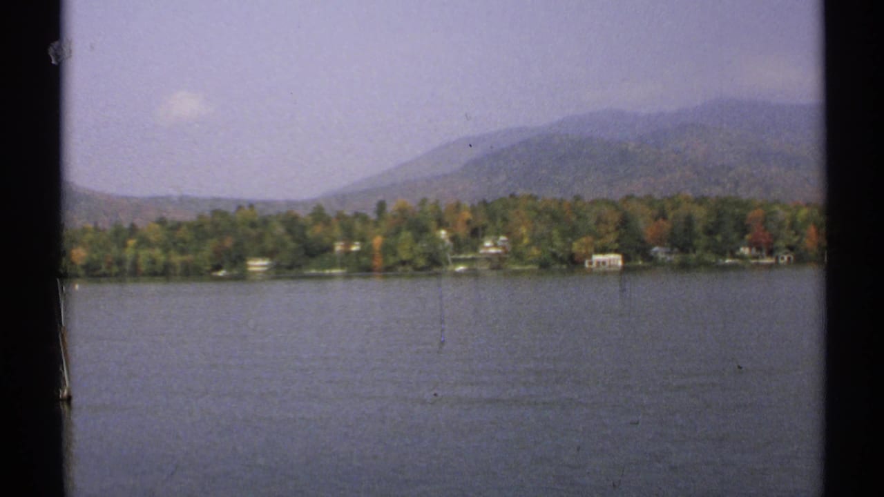 1971 New York frame showing lake water, small shoreline houses, autumn-colored trees, and distant mountains.