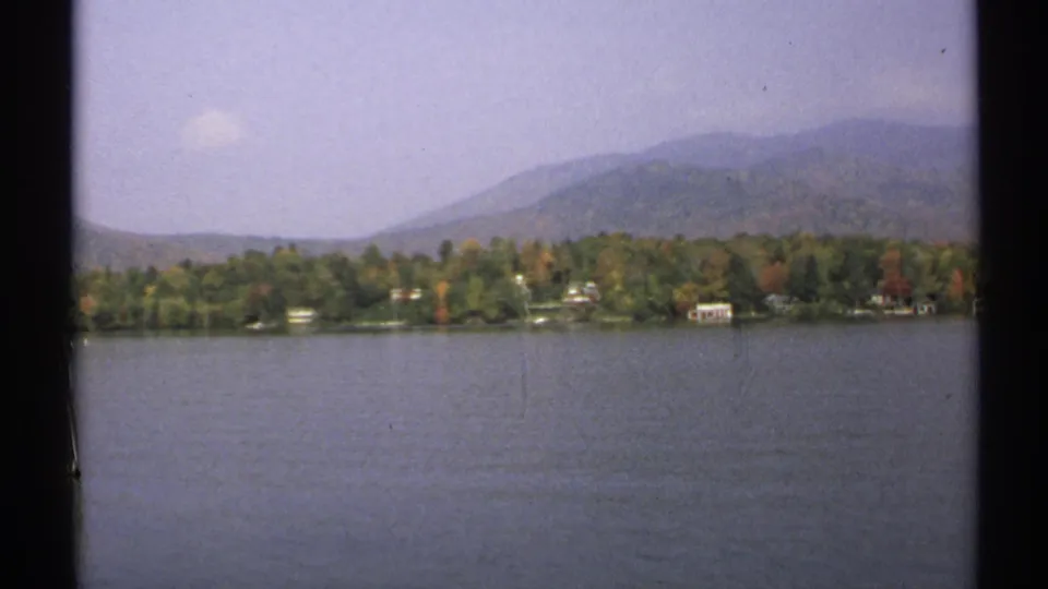 1971 New York 8mm view across a calm lake toward autumn trees, small shoreline houses, and hazy wooded mountains.