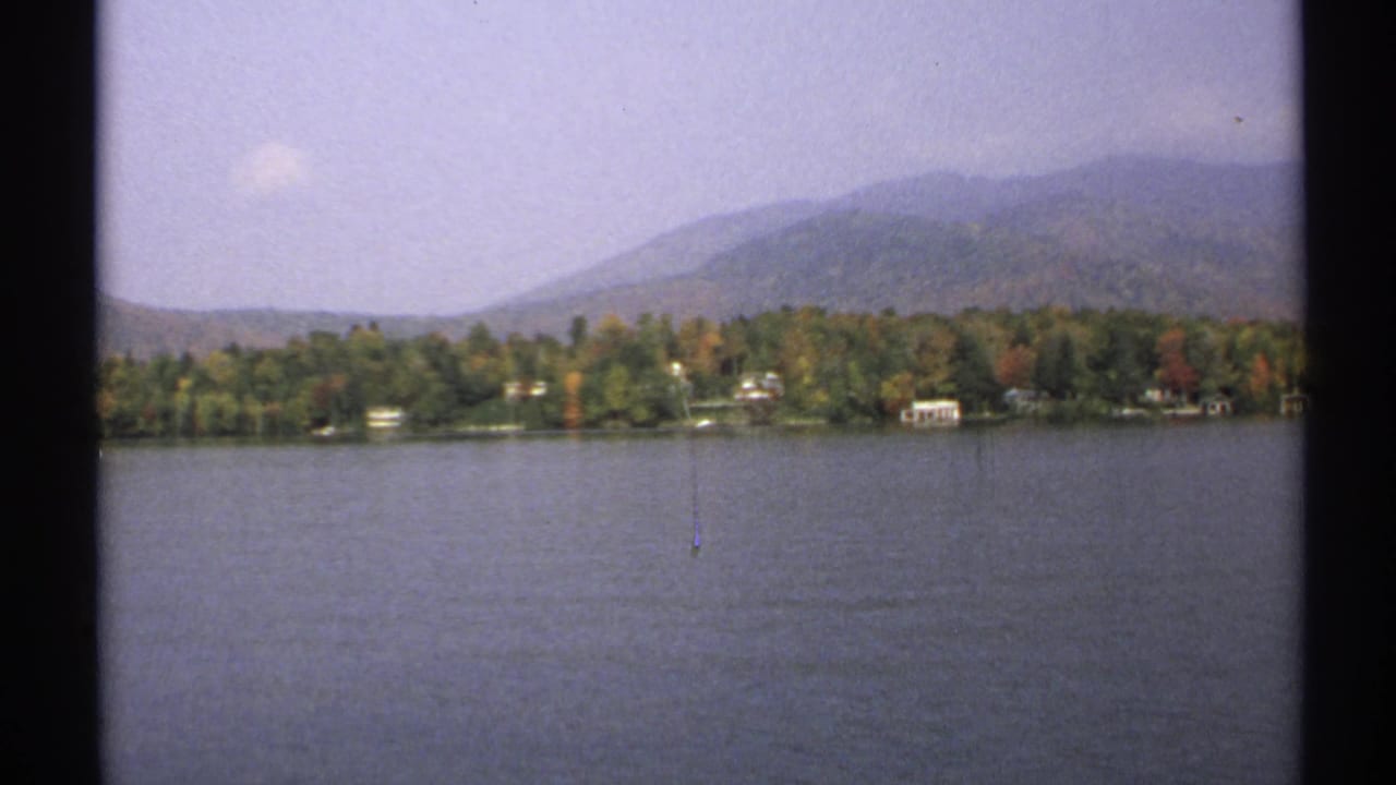 1971 New York lakeside home-movie frame showing muted fall foliage, scattered shoreline houses, and distant wooded hills under a pale sky.