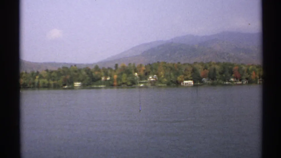 1971 New York lakeside home-movie frame showing muted fall foliage, scattered shoreline houses, and distant wooded hills under a pale sky.