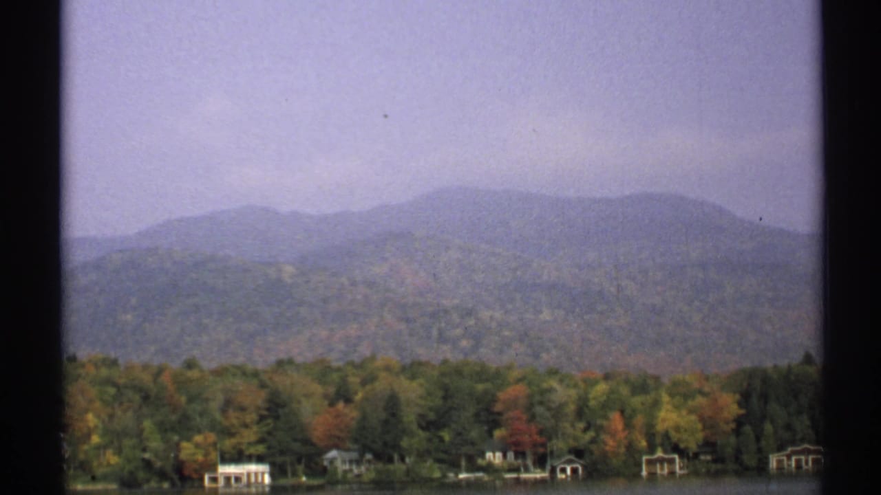 1971 New York 8mm frame with autumn treetops, a narrow shoreline, small boathouse-like buildings, and a hazy mountain ridge.