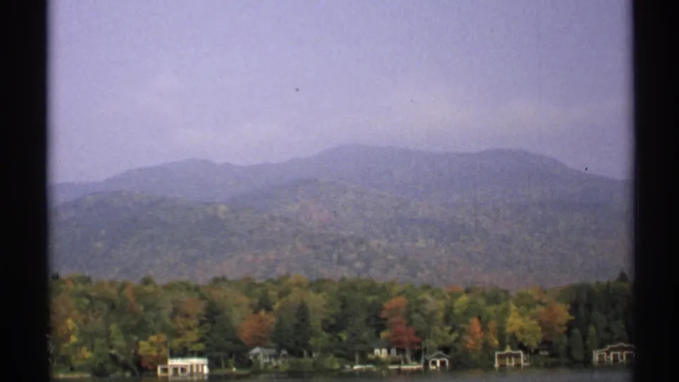 1971 New York 8mm frame with autumn treetops, a narrow shoreline, small boathouse-like buildings, and a hazy mountain ridge.